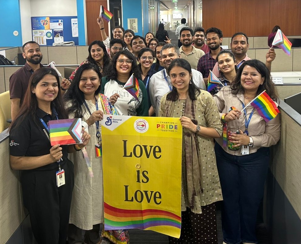 A group of cheerful people smiling and holding Rainbow flags, along with a banner that reads "Love is Love," featuring the GeneralMills logo in the top left corner. The scene depicts a celebration for the Mumbai Queer Azadi Month.