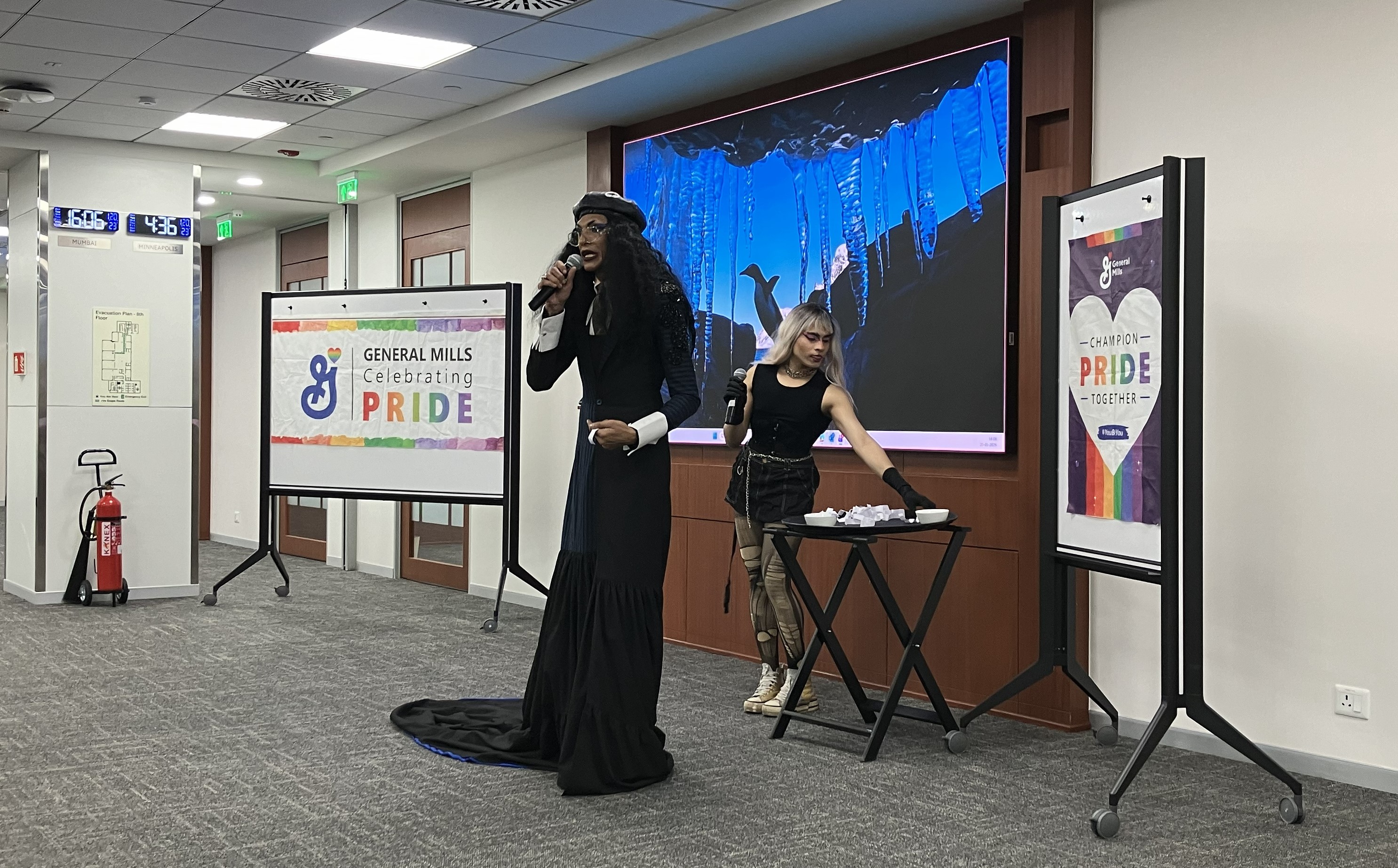 An indoor Pride-themed event is taking place in a conference room. A performer dressed in a long black outfit is holding a microphone and speaking on stage. Another person stands beside a small table with papers or cards arranged on it. Behind them is a large screen displaying an image of blue and black abstract shapes. On either side of the stage are standing boards with Pride-themed posters, including one that reads “General Mills Celebrating Pride.” The room has office lighting, carpeted flooring, and fire safety equipment visible in the background.